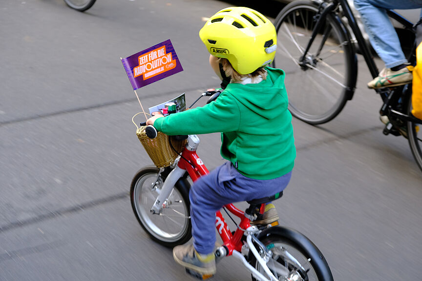 Kidical Mass Friedrichshain/Lichtenberg Ein Kind fährt auf einem roten Rad mit Radvolutionsflagge am Lenker.