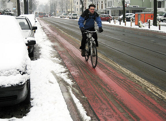 Ein Radfahrer in Winterkleidung fährt auf einem rot markierten Radfahrstreifen neben einer Straße, der weitgehend von Schnee befreit ist, während am Straßenrand und auf geparkten Autos Schnee liegt.